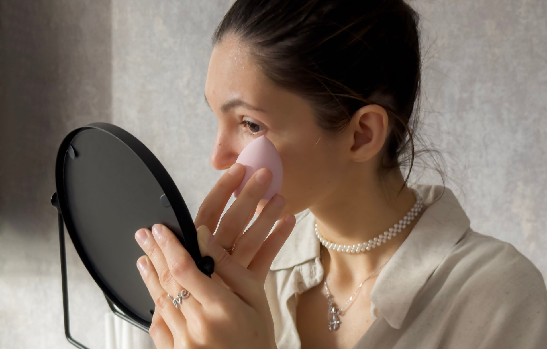A woman uses a sponge to blend foundation on her face while looking into a mirror, focusing on her makeup application in a bright setting